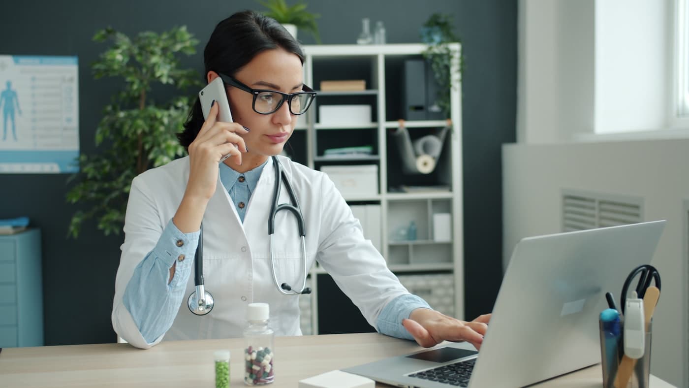 A physician recording a visit summary on her phone while reviewing the note on her laptop